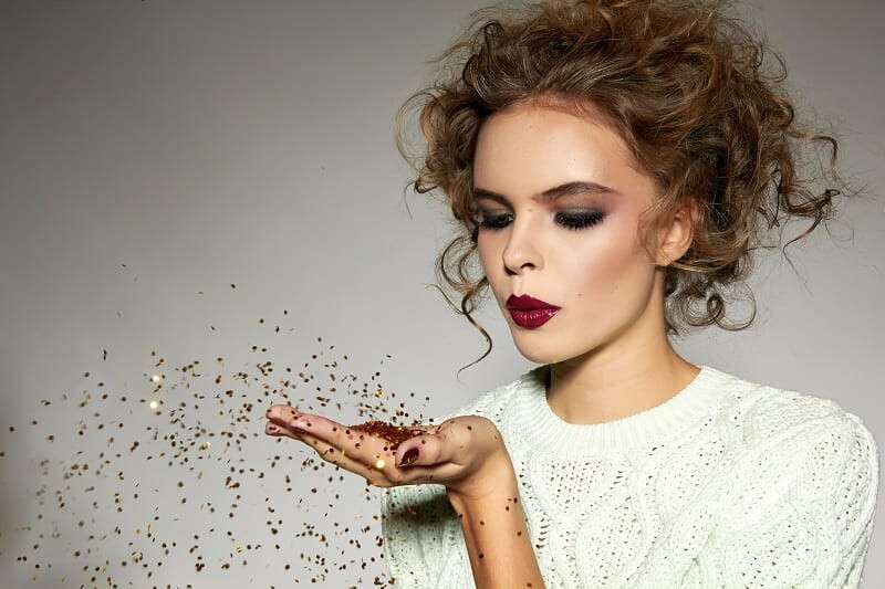 Beautiful sexy young blond girl with wavy hair bright evening make-up red lips long fluffy eyelashes holds palm with gold sequins in front of him and blows them a holiday New Year Christmas joy fun