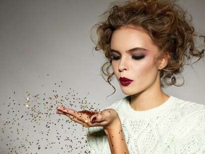 Beautiful sexy young blond girl with wavy hair bright evening make-up red lips long fluffy eyelashes holds palm with gold sequins in front of him and blows them a holiday New Year Christmas joy fun